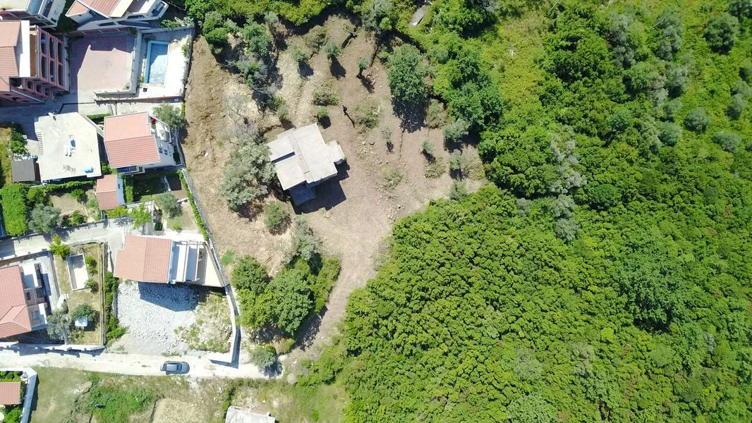 Aerial view of a plot of land with a small building, surrounded by dense forest and neighboring houses.