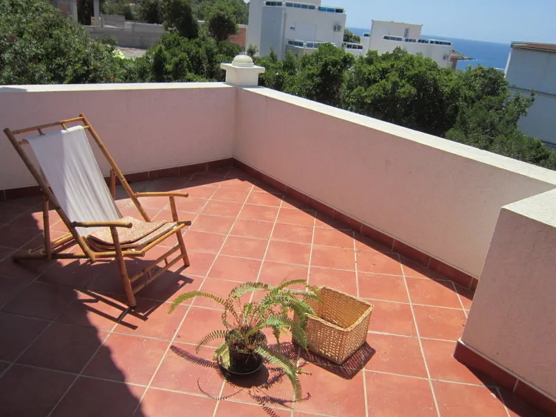 Terrace with terracotta tiles, wooden deck chair, potted plant, overlooking sea and trees.