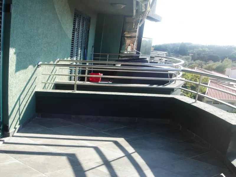Tiled terrace with metal railing overlooking hills and neighboring rooftops under bright sun.