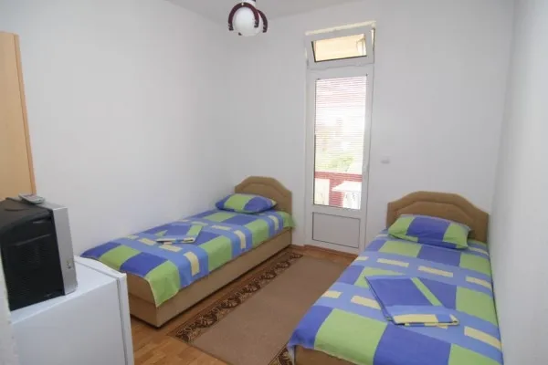 Two single beds with blue-green bedding, TV on a cabinet, window leading to a balcony, wooden floor.