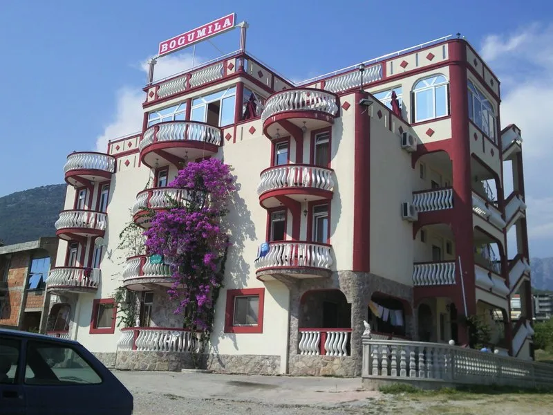 Exterior view of a multi-story building with curved balconies, red trim, and purple flowering vines on the facade under a blue sky.