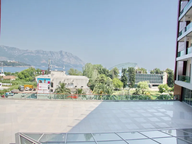 Wide tiled balcony with glass railing overlooking sea, mountains, and nearby buildings.