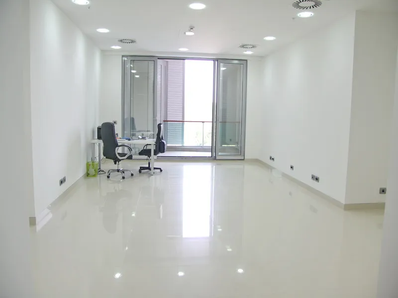 Empty office space with white walls, glossy floor, and a desk with two chairs near glass sliding doors leading to a balcony.
