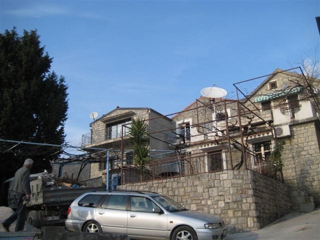 Two-story stone house in Rafailovici, Montenegro, with a terrace, grapevine, and a parked car.