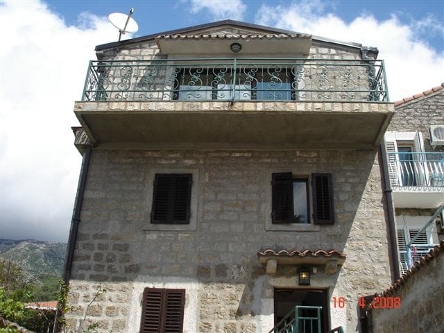 Facade of a stone house in Rafailovici, Montenegro, with a wrought-iron balcony, tiled roof, and window shutters.
