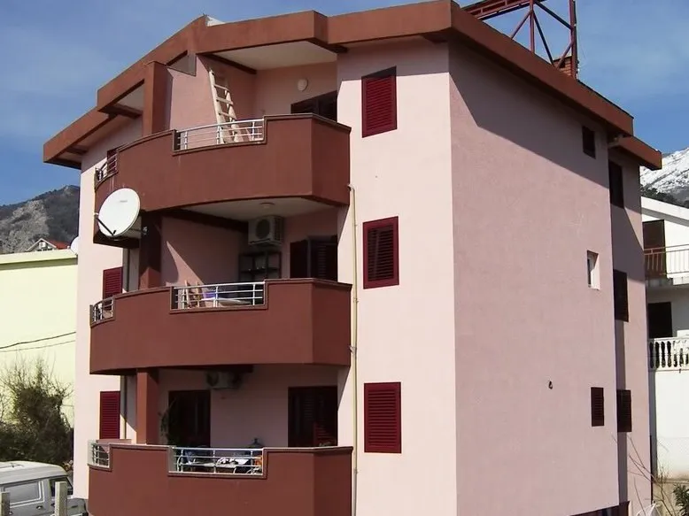 Exterior view of a multi-story residential building featuring balconies, red shutters, and a satellite dish against mountains.