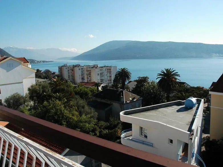 View from a balcony overlooking the sea, mountains, and city buildings. A railing is visible in the foreground.