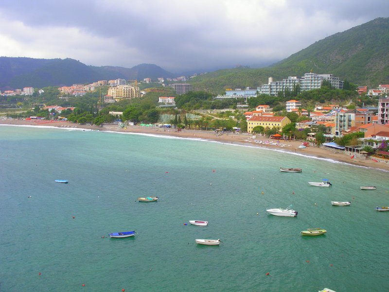 Panoramic view of the sea, beach, and mountains in the resort village of Rafailovići, Montenegro, with boats in turquoise water.