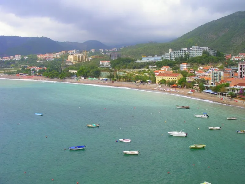 Panoramic view of a sea bay with boats, sandy beach, and coastal buildings set against green mountains.