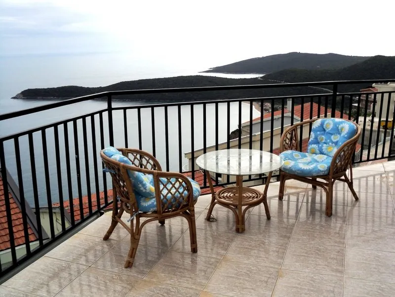 Balcony with two wicker chairs and a glass table overlooking the sea and hills.