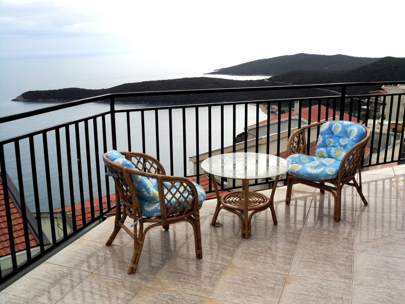 Villa balcony in Budva, Montenegro: wicker furniture, a glass table, and panoramic views of the sea and mountains.