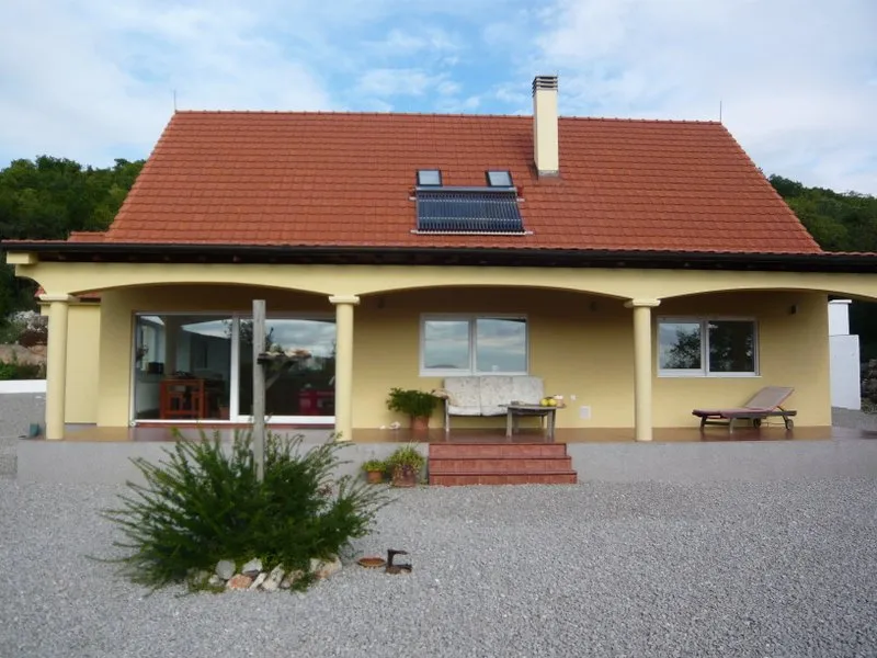 Exterior view of a single-story house with a red tiled roof, covered terrace, and gravel yard.