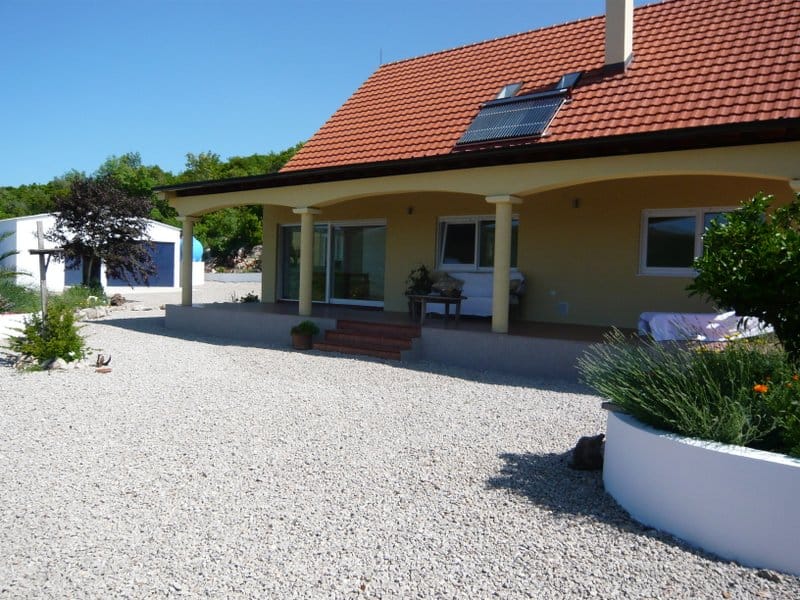 Single-story house with a terrace, tiled roof, and solar panels in Nikšić, Montenegro. Gravel-covered lot.