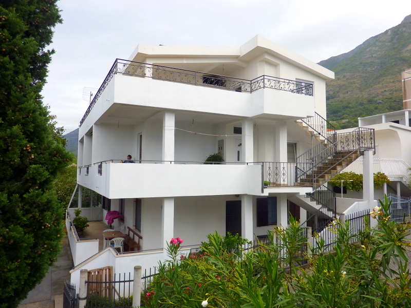Three-story white house with terraces and an external staircase in Sutomore, Montenegro, surrounded by greenery and mountains.
