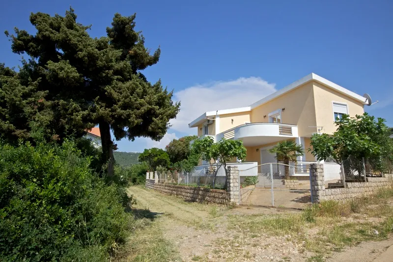 Exterior view of a two-story yellow house with a balcony, surrounded by trees and a stone wall under a blue sky.