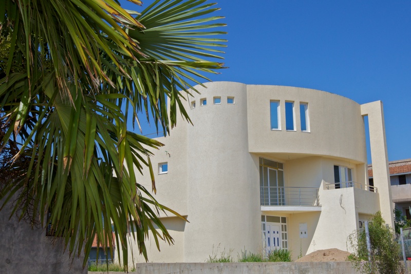 Modern two-story house with panoramic windows and a balcony in Utjeha, Montenegro, with a palm tree and blue sky in the background.