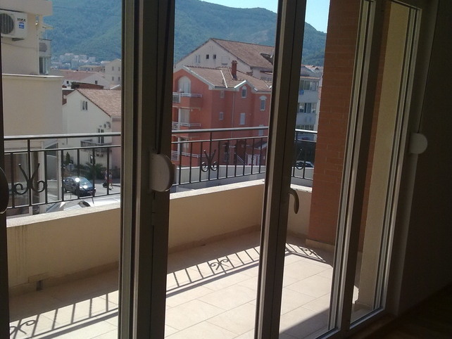 View from the apartment balcony in Budva, Montenegro: panoramic windows, a tiled terrace, and a view of city buildings and mountains.