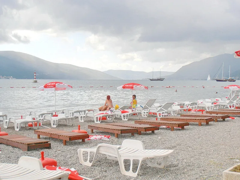 Gravel beach with white sunbeds and red umbrellas overlooking the sea, mountains, and sailboats.