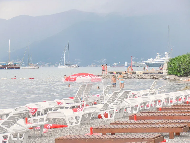 Peppered beach with white sun loungers and umbrellas near water, mountains and boats in background.