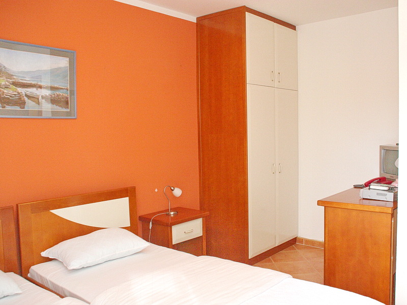 Bedroom in a hotel in Tivat, Montenegro: two beds, a wooden wardrobe, a bedside table, and an orange wall with a painting.