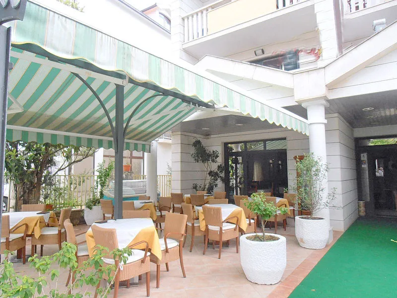 Outdoor terrace with dining tables under a striped awning, wicker furniture, white stone columns, and potted plants.