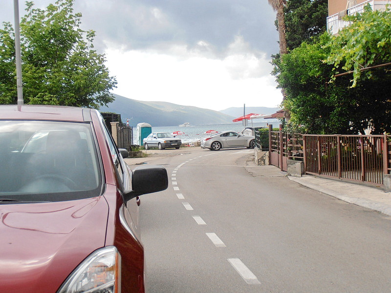 View of the road to the sea and the beach near the hotel in Tivat, Montenegro, with parked cars and green trees.