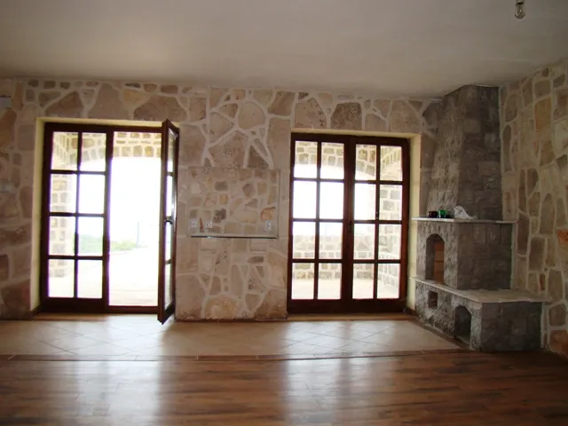 Empty room with stone walls, wood floor, and a large stone fireplace next to glass doors.