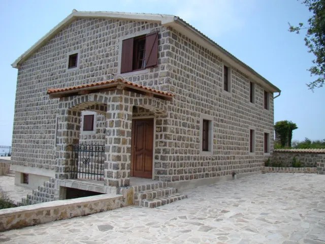 Stone house exterior with wooden door and porch, showing masonry walls and roof under sky.
