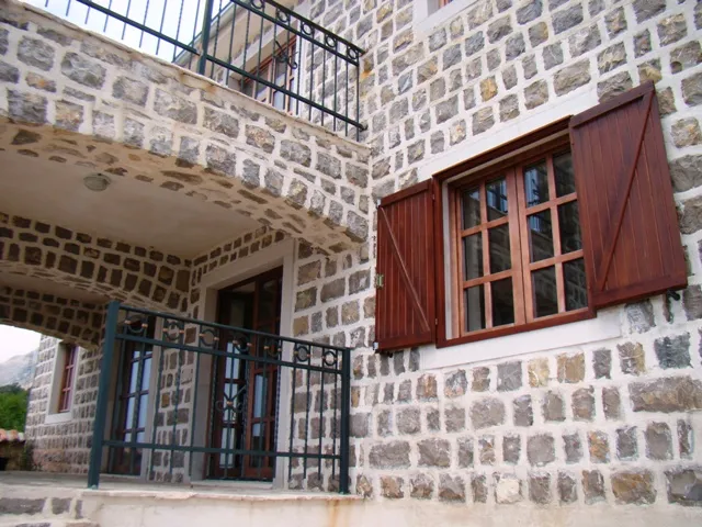Stone building facade featuring wooden windows with shutters, an upper balcony with iron railing, and a ground-level archway.