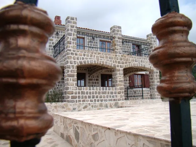 View of a stone house with balconies through a wrought iron fence. Grey stone facade, terrace and windows visible.
