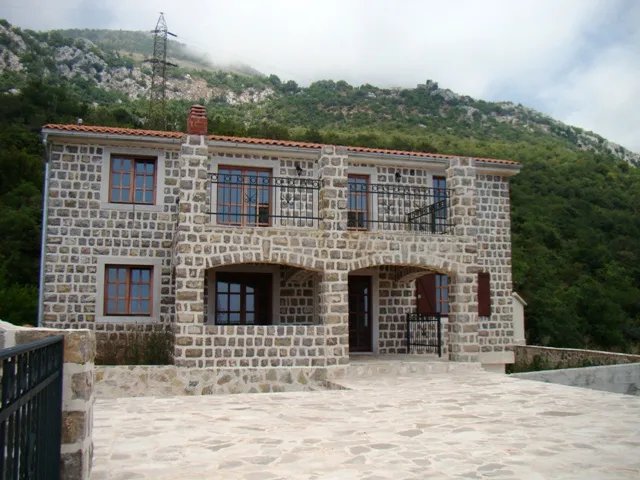 Exterior view of a two-story stone house with a balcony against a forested mountain backdrop.