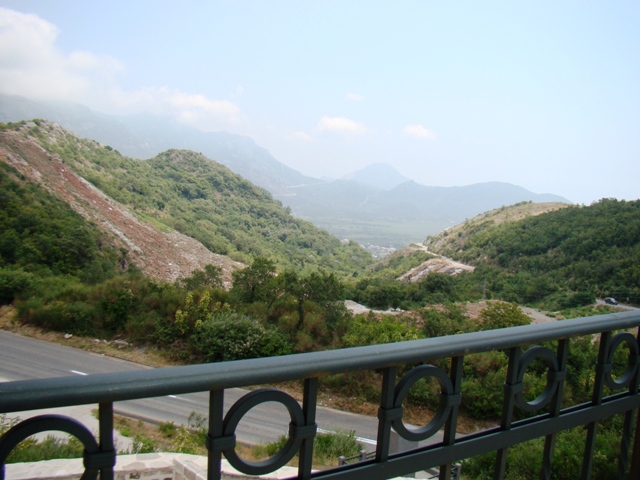 View from the balcony of a house in Petrovac, Montenegro: a panorama of green mountains and a mountain road with a wrought-iron railing.