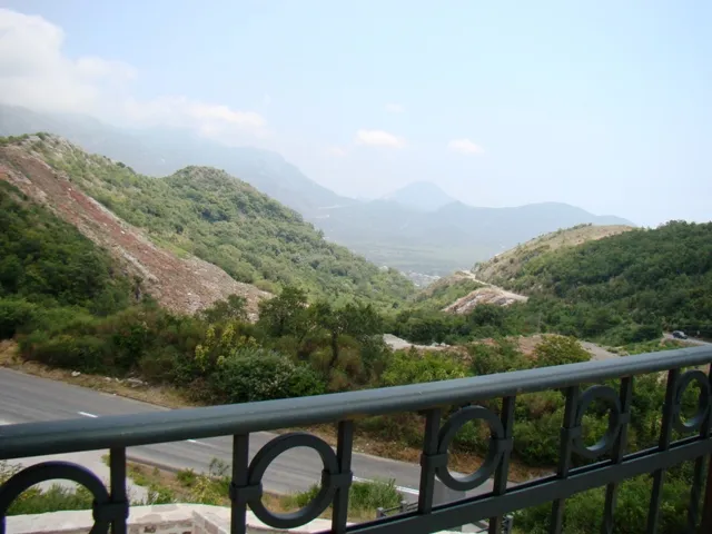 Mountain and road view from a balcony with a metal railing visible in the foreground.