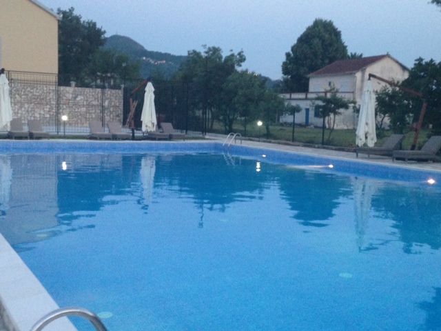 Outdoor swimming pool with sun loungers and umbrellas at a residential complex in Budva, Montenegro, with mountain views and green grounds.