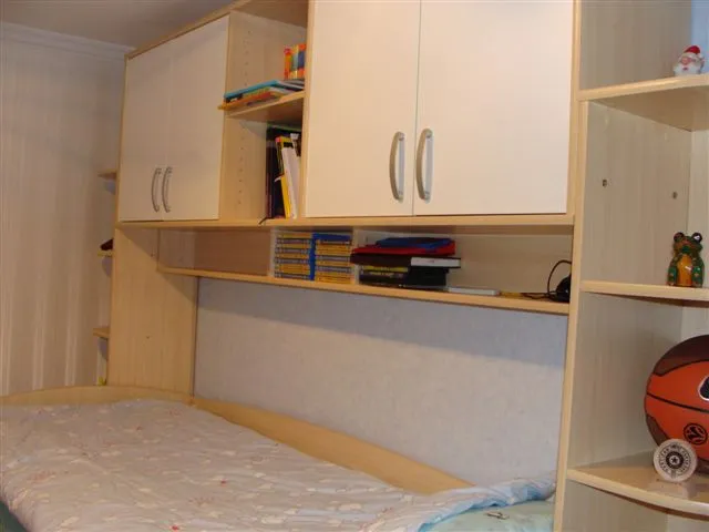 Bed with headboard and built-in light cabinets containing books, discs, and toys on shelves.