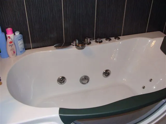 White jacuzzi bathtub with chrome jets against dark tiled wall. Toiletries bottles are visible on the left edge of the tub deck.