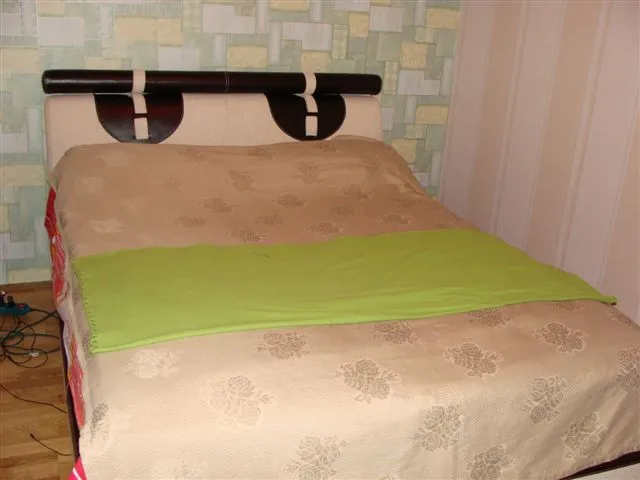 Bed with beige cover and green blanket against a wall with geometric wallpaper.