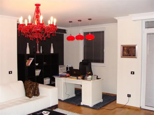 Living room featuring a white sofa, black shelving unit, and red chandelier. A home office desk with a laptop is also visible.