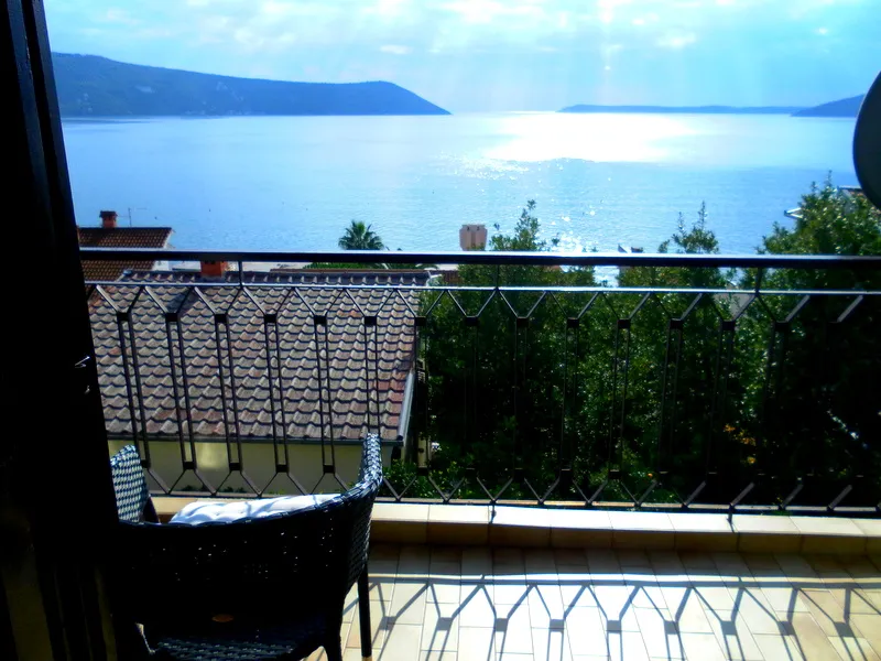 Balcony with black wicker chair and metal railing overlooking the sea and tiled rooftops.