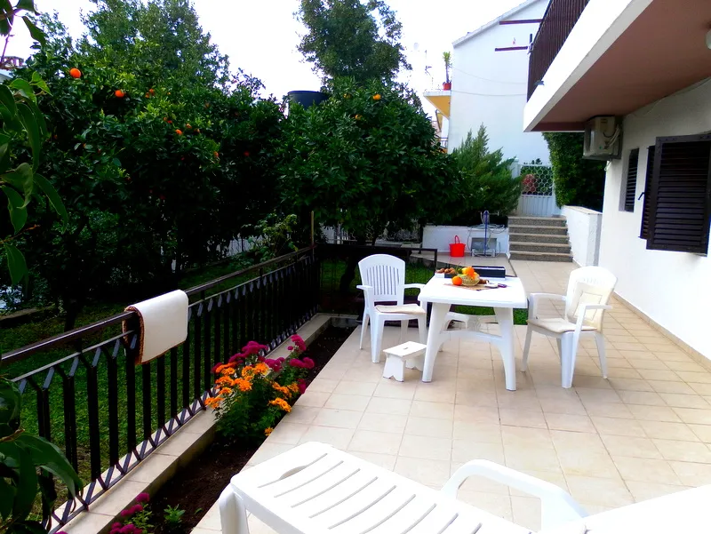 Tiled terrace with white plastic furniture, black railing, and orange trees next to a white house exterior.