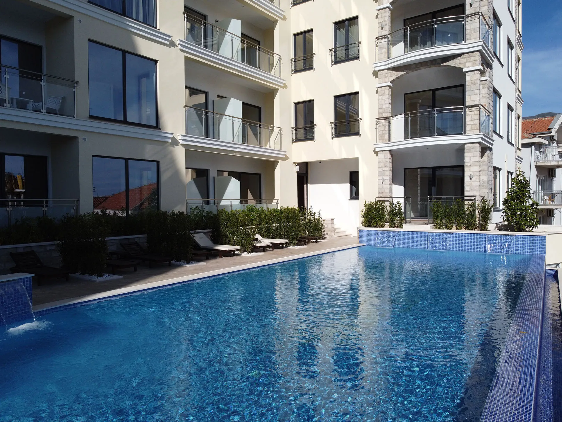 Large blue tiled swimming pool with a small waterfall feature in front of a multi-story residential building with balconies.