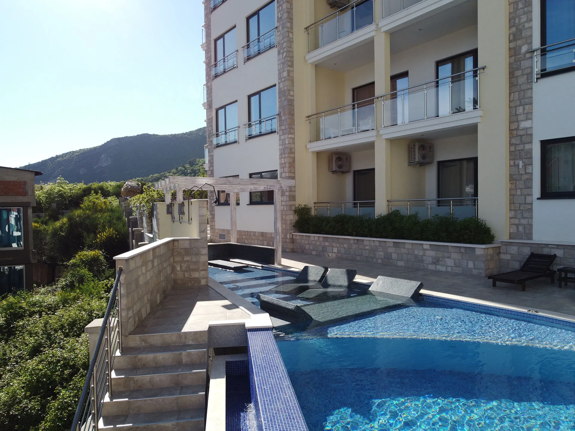Outdoor swimming pool with blue tiles, terrace with sunbeds, and a multi-story building against mountains.