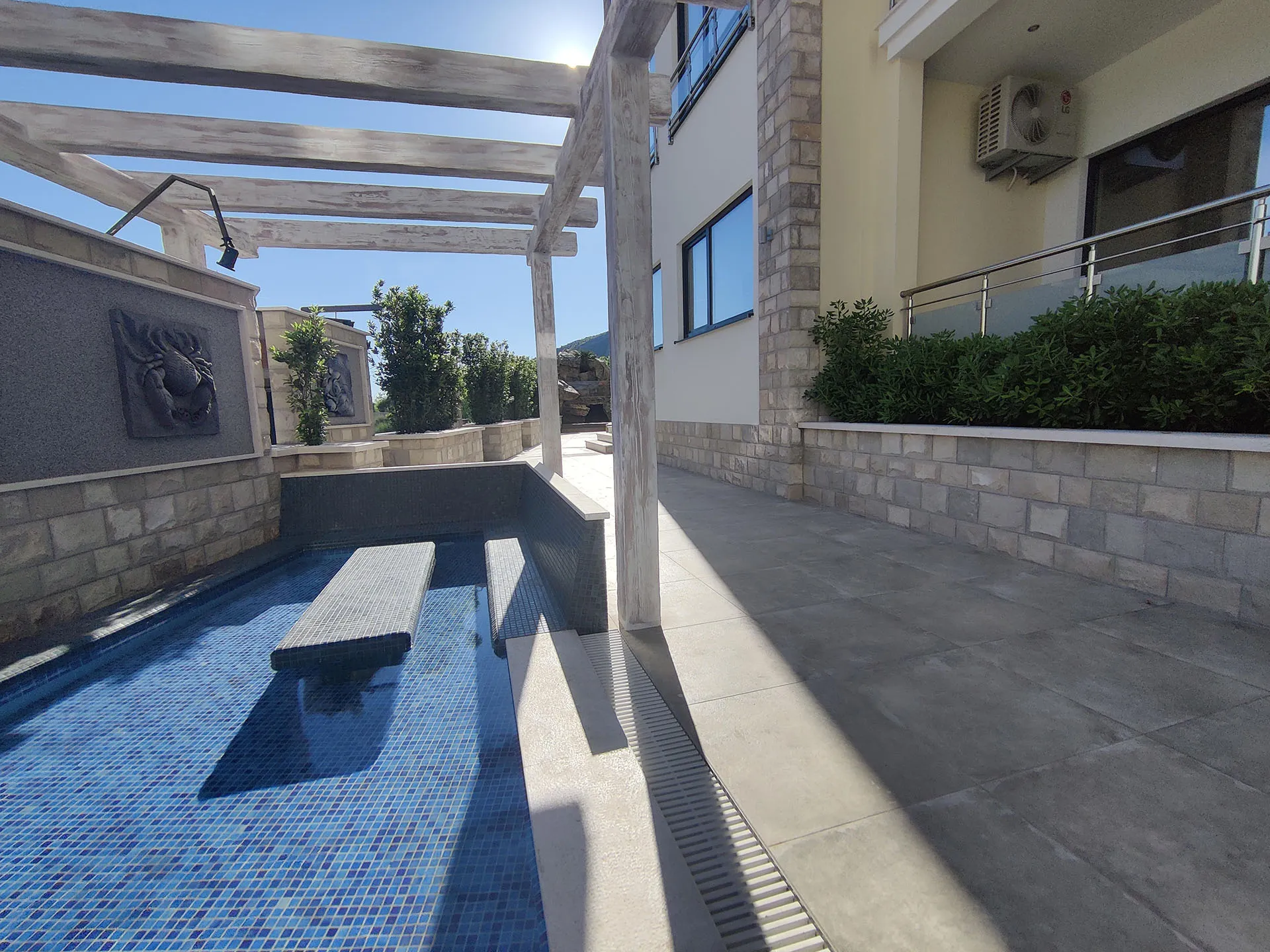 Small blue mosaic pool with wooden pergola on an open terrace next to a building facade.