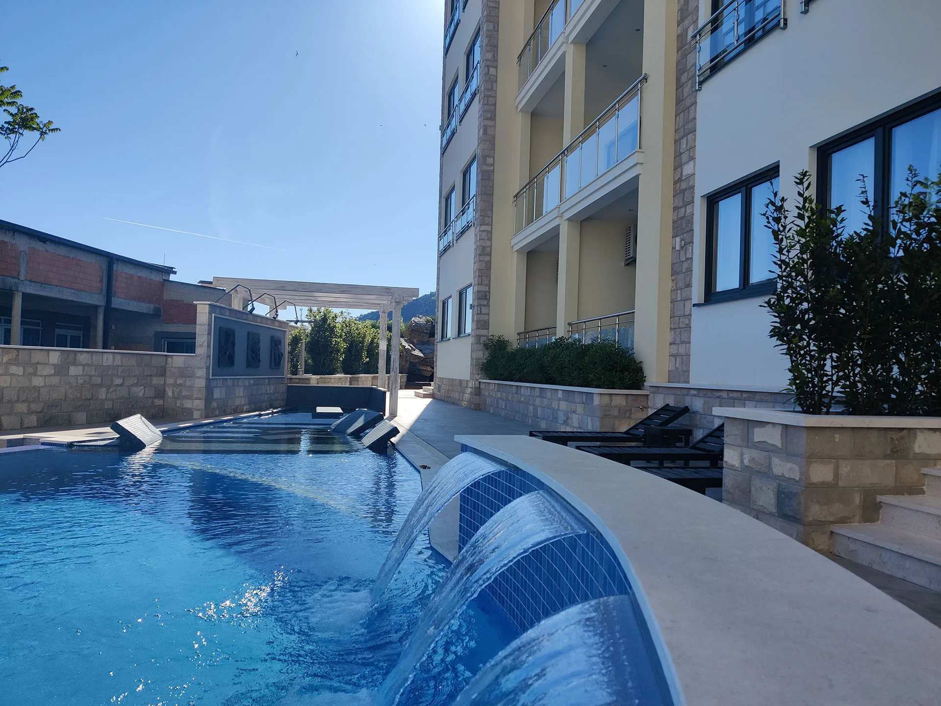 Outdoor swimming pool with waterfalls and blue tiles, located next to a multi-story building and sun loungers.