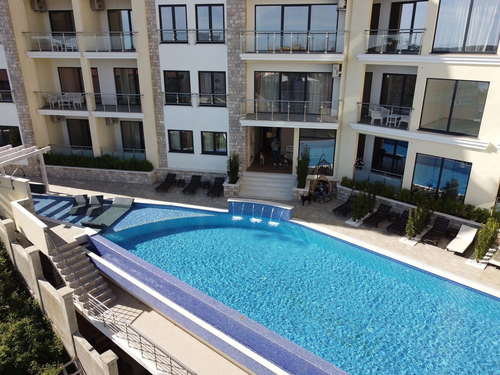High-angle view of a large blue swimming pool with water jets and sunbeds in front of a multi-story building.