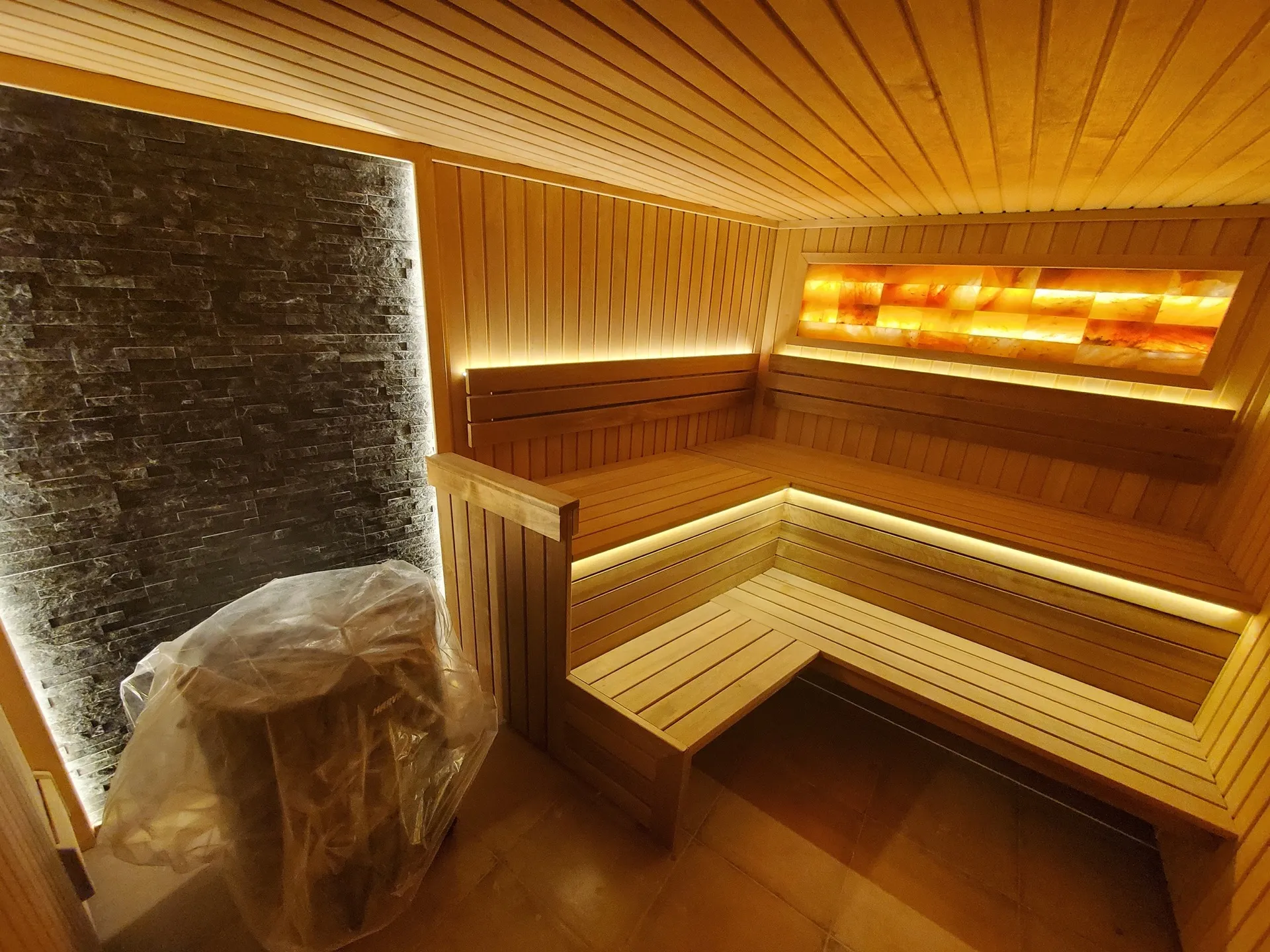 Interior of a sauna featuring wooden benches, warm LED lighting, a salt lamp panel, and a stone wall section.