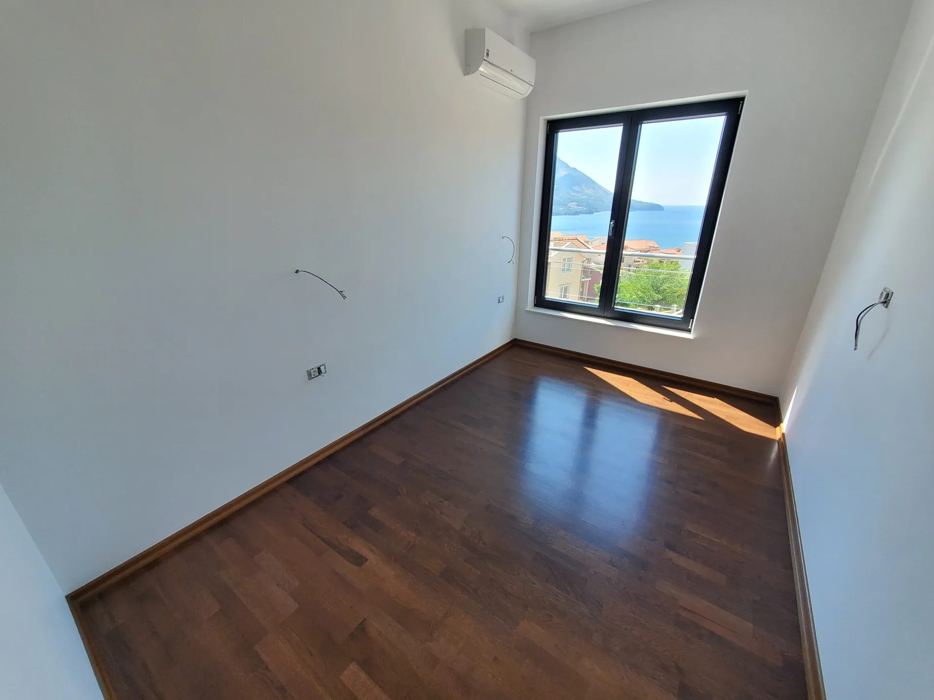 Empty room with dark wood flooring, wall-mounted AC unit, and a window overlooking the sea and mountains.