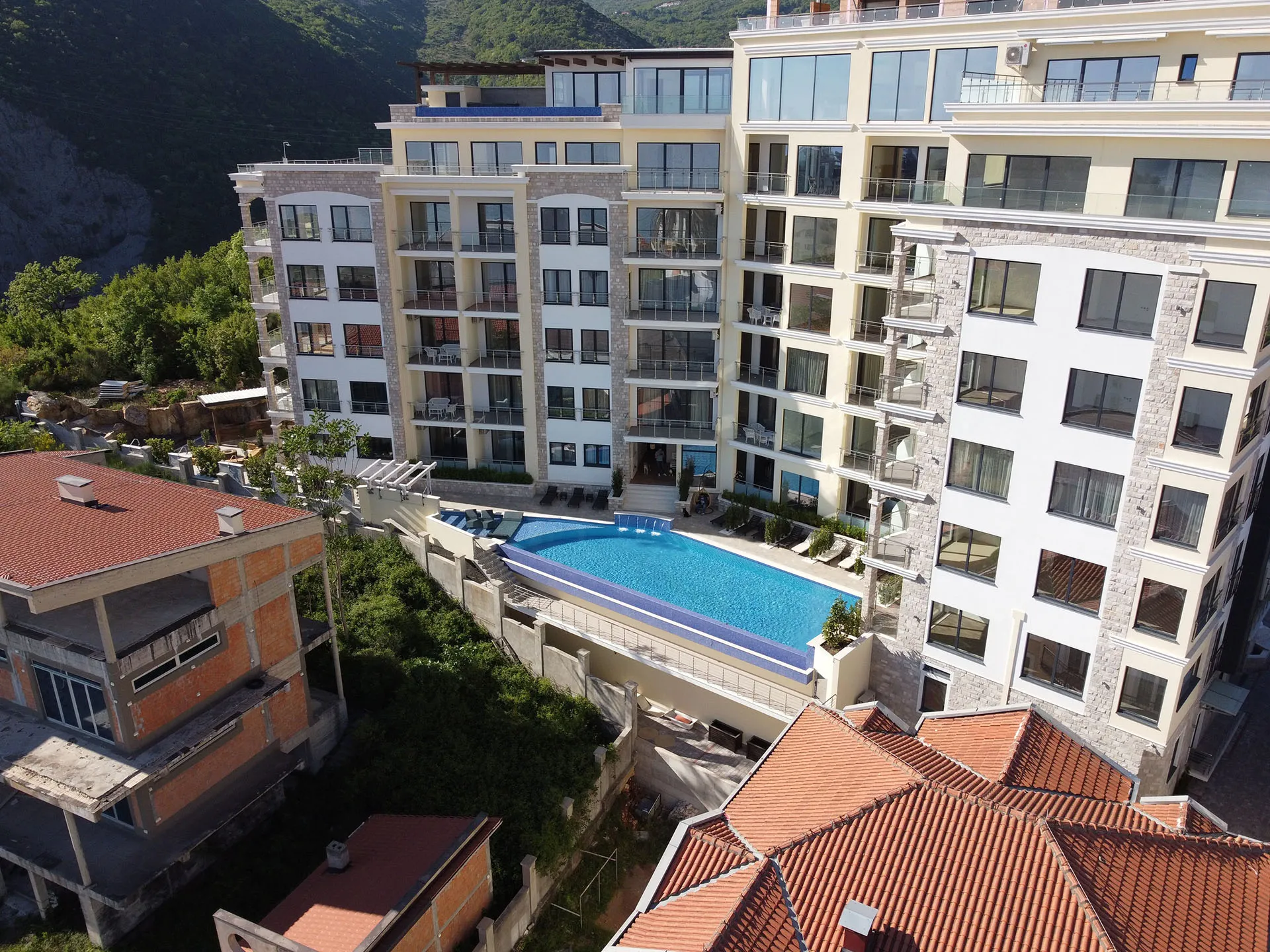 Aerial view of a multi-story residential complex featuring a large blue swimming pool and lounge chairs against mountains.