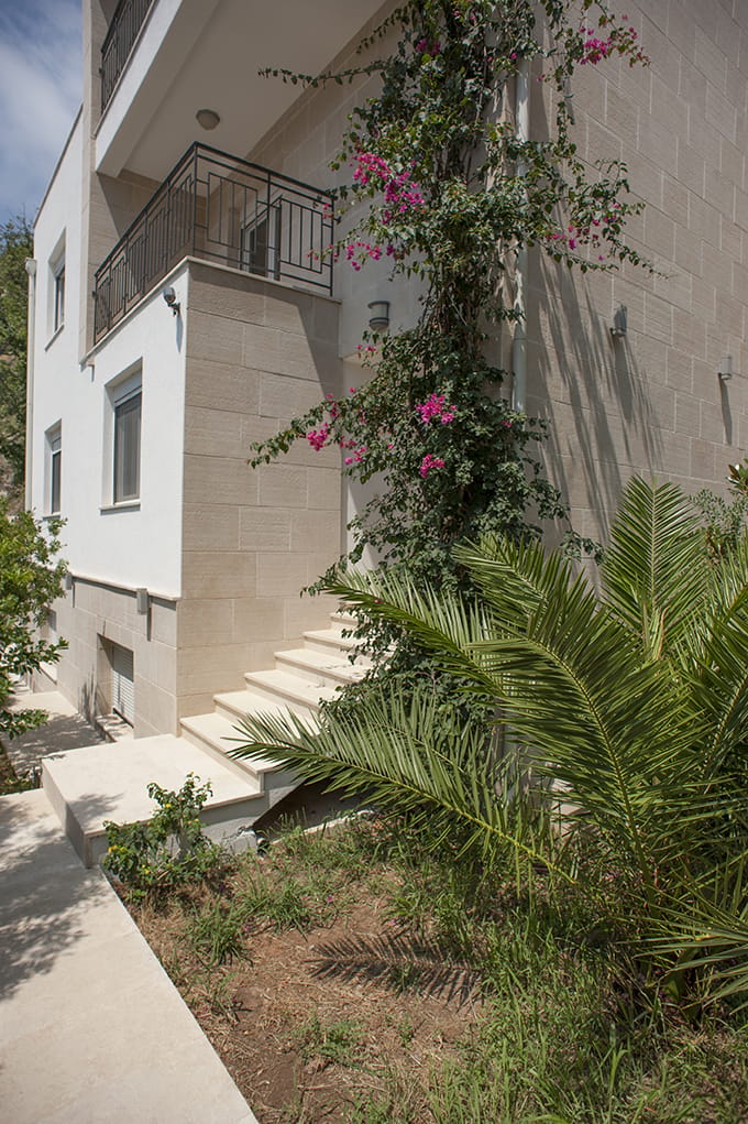 Facade of a modern villa in Perazica Do, Montenegro: stone cladding, balcony, blooming bougainvillea, and a palm tree by the entrance.