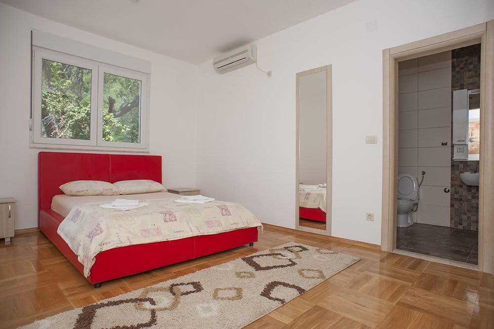 Bedroom with a red bed and en-suite bathroom in a villa in Perazica Do, Montenegro: parquet flooring, air conditioning, and garden view.